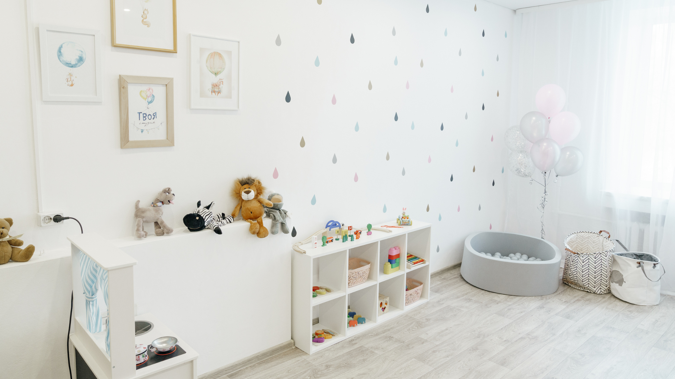 A Bright and airy playroom with a play kitchen, a ball bin, some stuffed animals and 12 toys on a shelf.